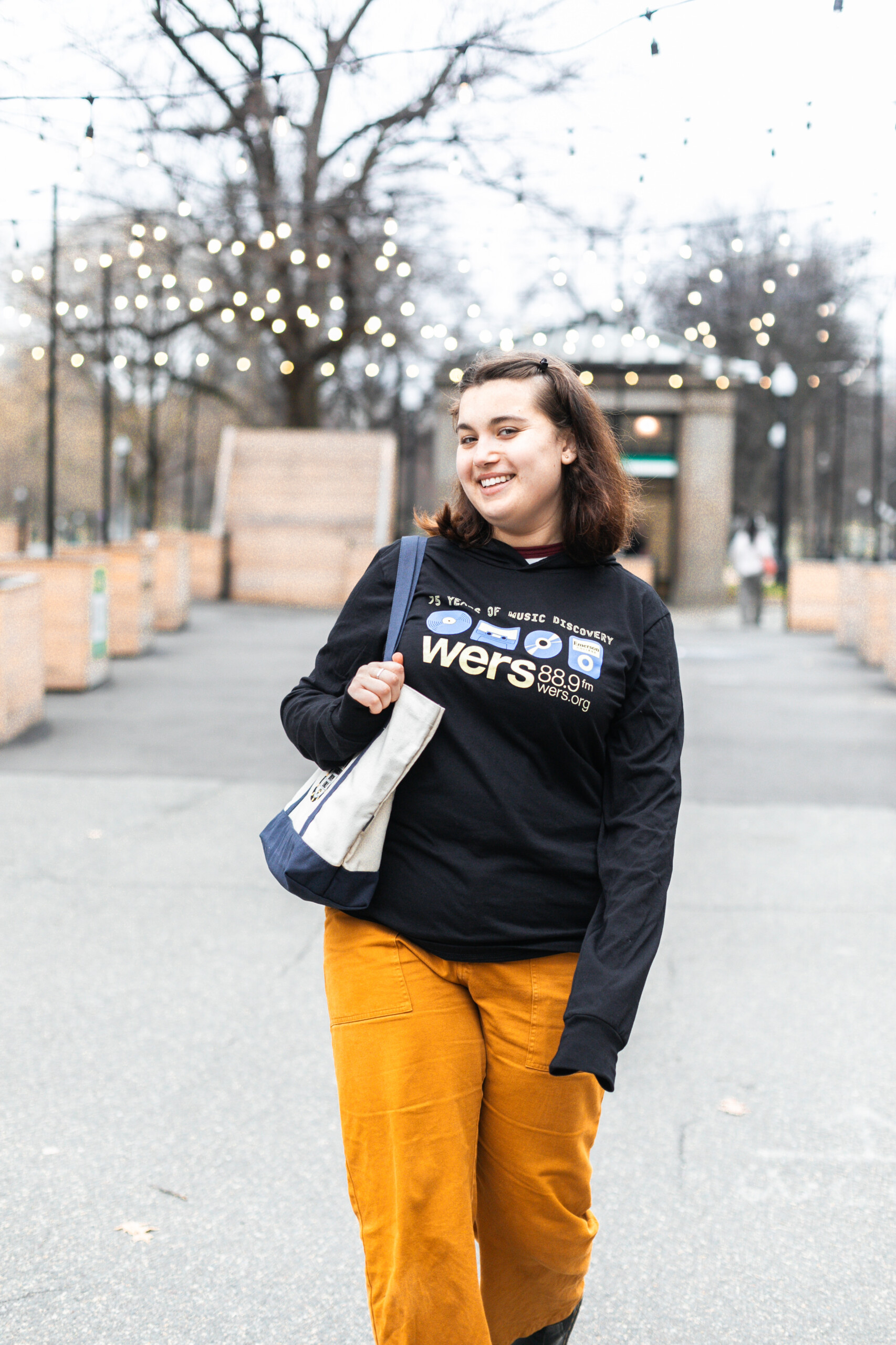 Photo of Emelia, host of Standing Room Only, wearing a black long-sleeved shirt with a "75 Years of Music Discovery, WERS 88.9" logo. She is also holding a cream tote with dark blue straps and an embroidered SRO logo in the center.