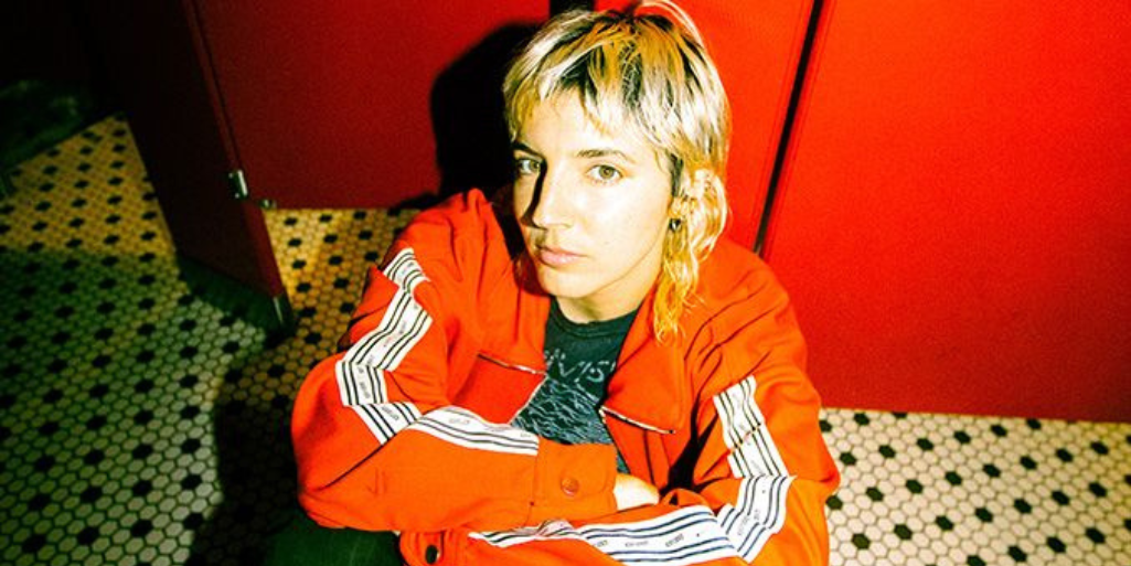 Caroline Rose sits against the door of a red bathroom stall on a black and white hexagon tiled floor. Rose is wearing a bright orange jacket and green pants.