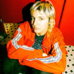 Caroline Rose sits against the door of a red bathroom stall on a black and white hexagon tiled floor. Rose is wearing a bright orange jacket and green pants.