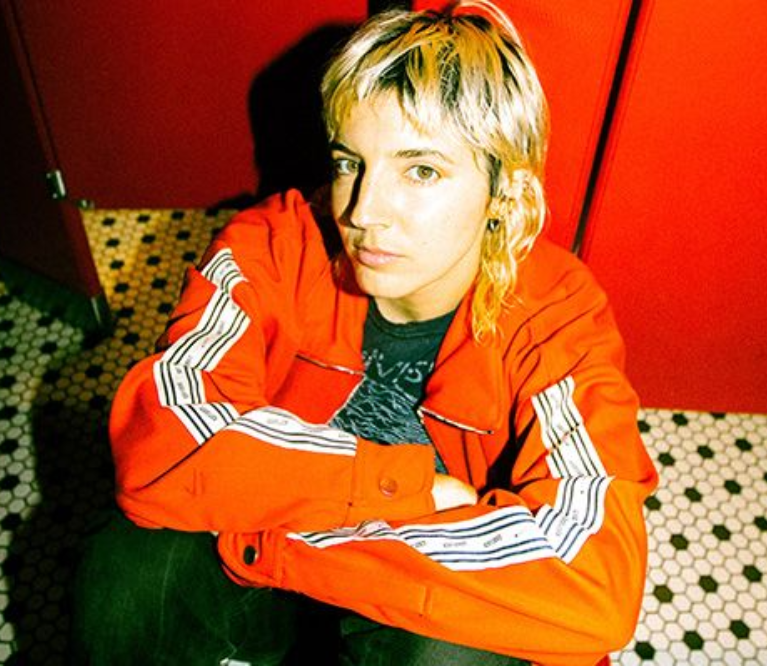 Caroline Rose sits against the door of a red bathroom stall on a black and white hexagon tiled floor. Rose is wearing a bright orange jacket and green pants.