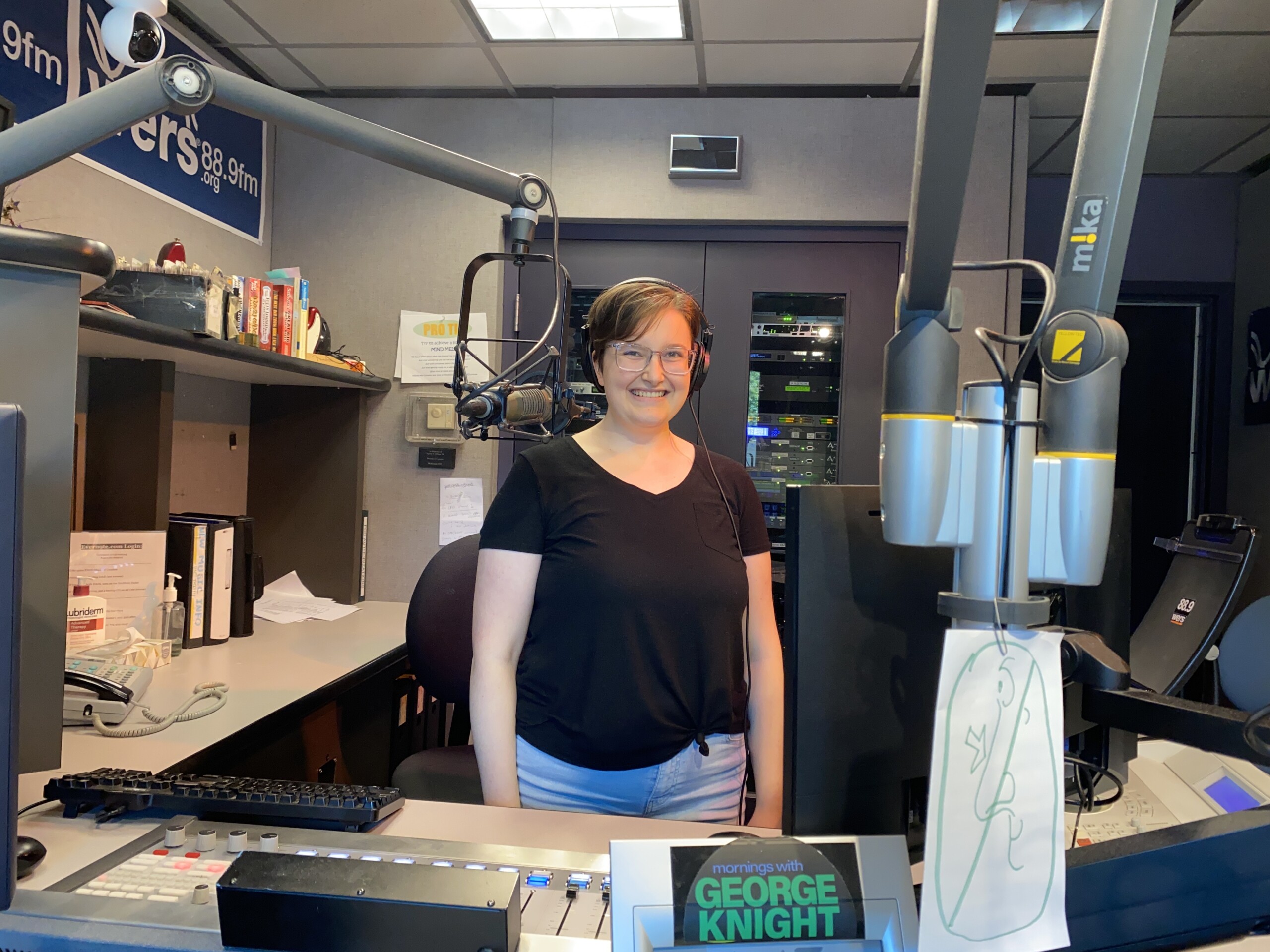 Picture of Eva smiling in the air studio. She is a white woman with short brown hair. She is wearing a black T-shirt and jeans.