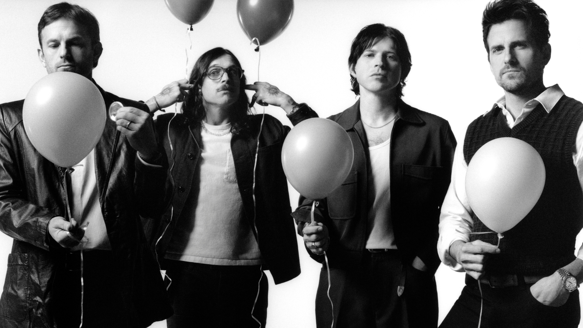 A black and white image of the members of Kings of Leon, four white men with brown hair, black pants, and black and white shirt and jacket combos. They are holding balloons but have serious facial expressions.