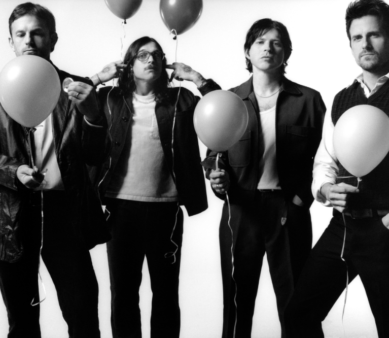 A black and white image of the members of Kings of Leon, four white men with brown hair, black pants, and black and white shirt and jacket combos. They are holding balloons but have serious facial expressions.
