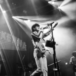 Declan McKenna in balck and white singing behind stage lights while holding his electric guitar.