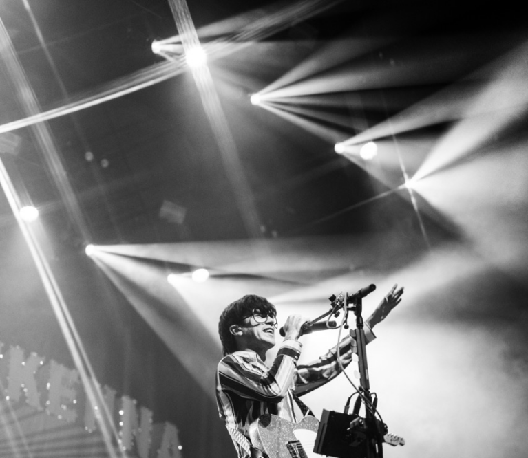 Declan McKenna in balck and white singing behind stage lights while holding his electric guitar.