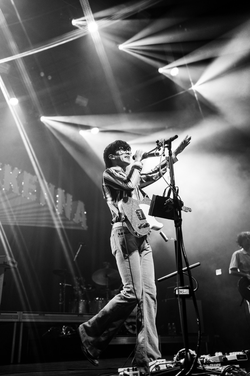 Declan McKenna in balck and white singing behind stage lights while holding his electric guitar.