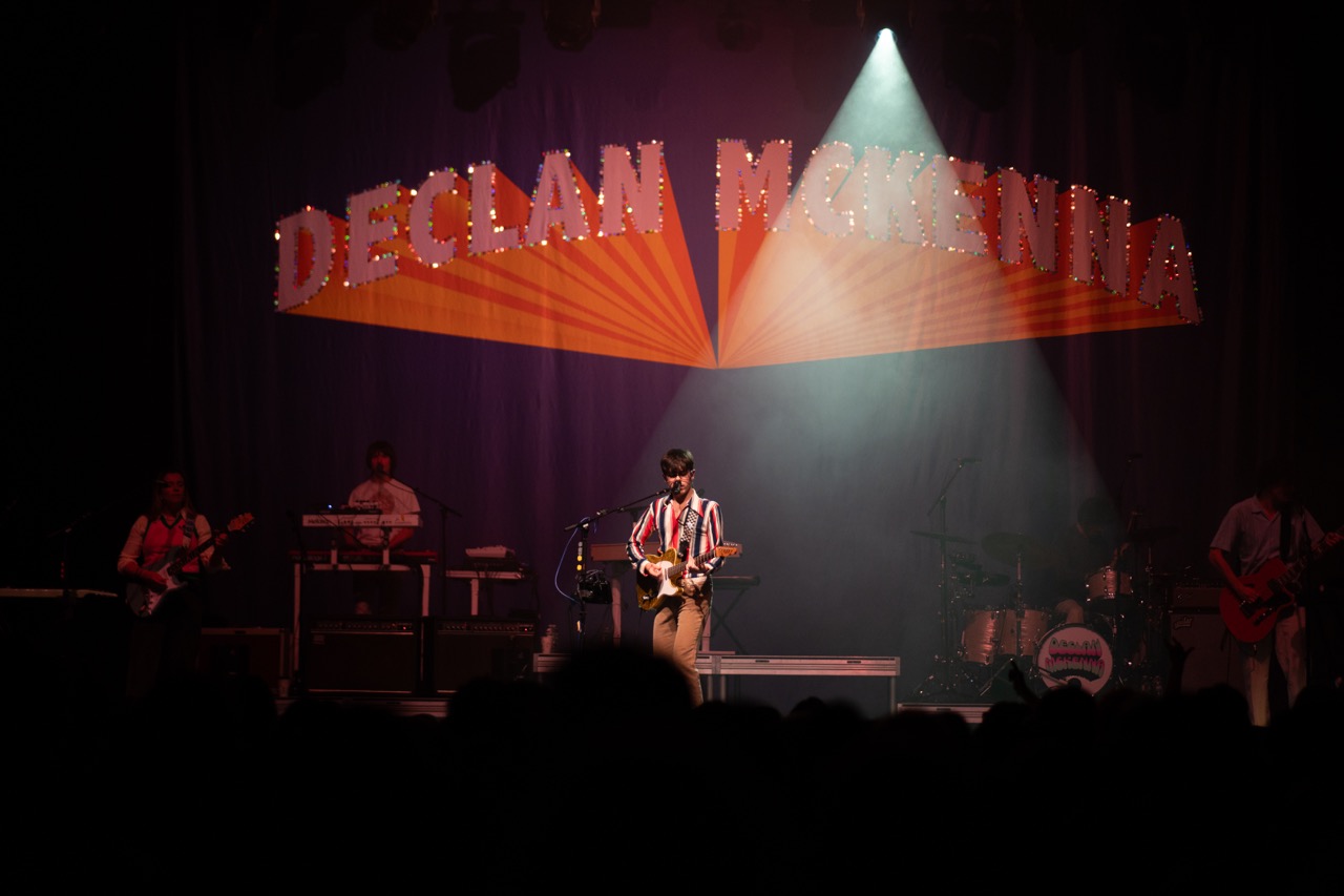 Photo of Declan McKenna on stage with a large spotlight on him while he plays guitar. Behind him a curtain reads "Declan McKenna" in red and orange 3-D style text.