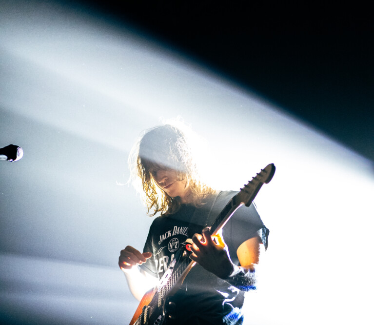 Mk.gee with a stage light behind him, strumming an electric guitar upwards.