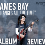 A photo of James Bay singing with his guitar while close-up images of him are translucent behind him on a grey-blue background. White text reads, "James Bay," Changes All the Time," "Album Review."