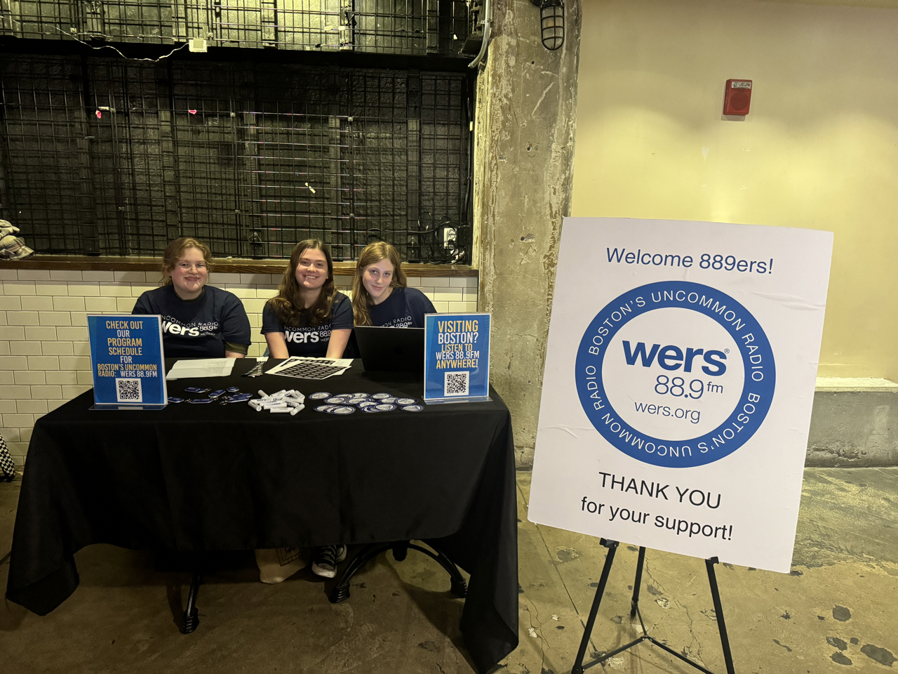 Three WERS employees are at a booth with swag. A white board with the circular WERS logo is in front of them.