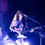 SASAMI strums a large, pointy, electric guitar onstage. She stands in front of light purple lighting.