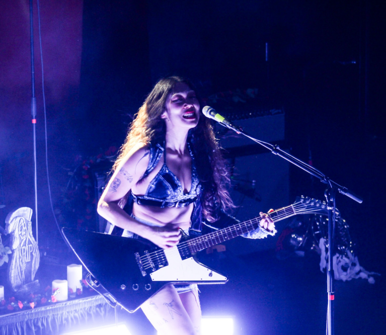 SASAMI strums a large, pointy, electric guitar onstage. She stands in front of light purple lighting.