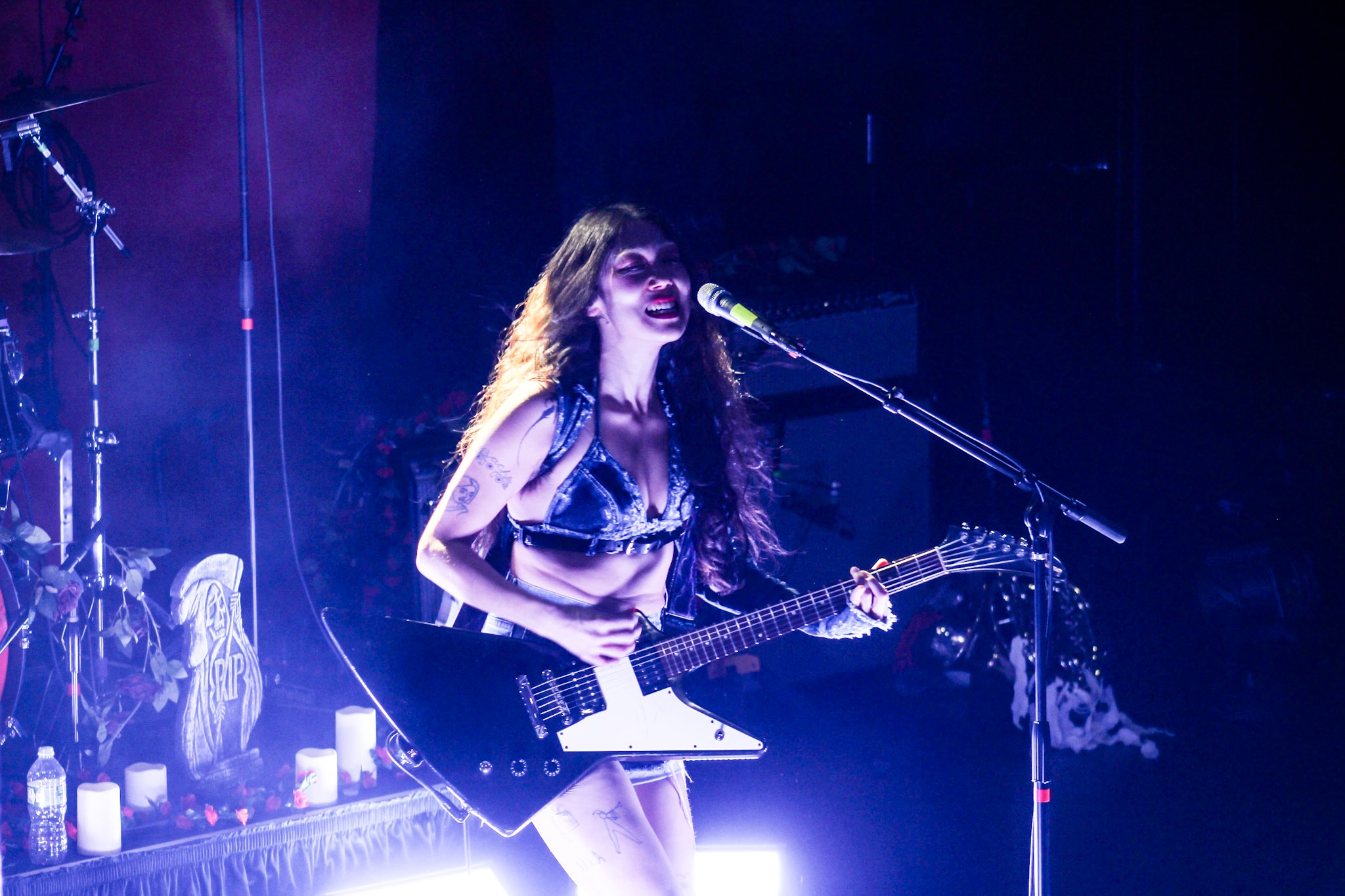 SASAMI strums a large, pointy, electric guitar onstage. She stands in front of light purple lighting.