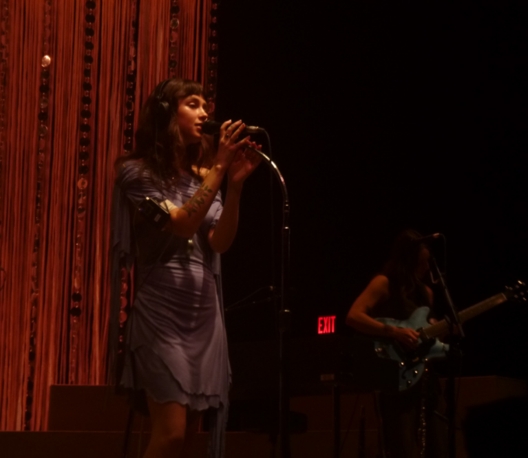 Clairo, onstage, singing in front of a red curtain.