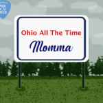 A roadside sign in front of an evergreen forest reads, "Ohio All the Time" in red and "Momma" in blue.