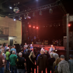 The Pixies play on the floor in front of a stage. Dozens of donors watch closely as red lights shine from the stage.