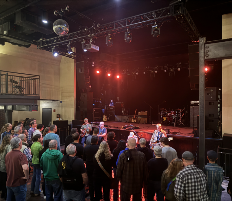 The Pixies play on the floor in front of a stage. Dozens of donors watch closely as red lights shine from the stage.