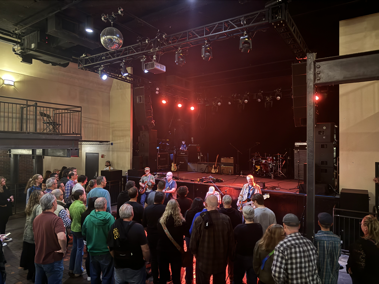 The Pixies play on the floor in front of a stage. Dozens of donors watch closely as red lights shine from the stage.