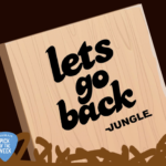 A wood panel has "Let's Go Back" and "Jungle" painted on it in black. The panel is behind short brown grass.