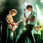 Two Hippo Campus Guitarists play while facing each other under light green lighting