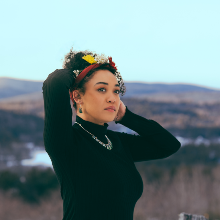 Farayi Malek, in a black turtleneck, put her hans on her curly hair in front of rolling hills.