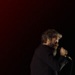 Father John Misty lowers his head in a medium shot while holding the microphone.