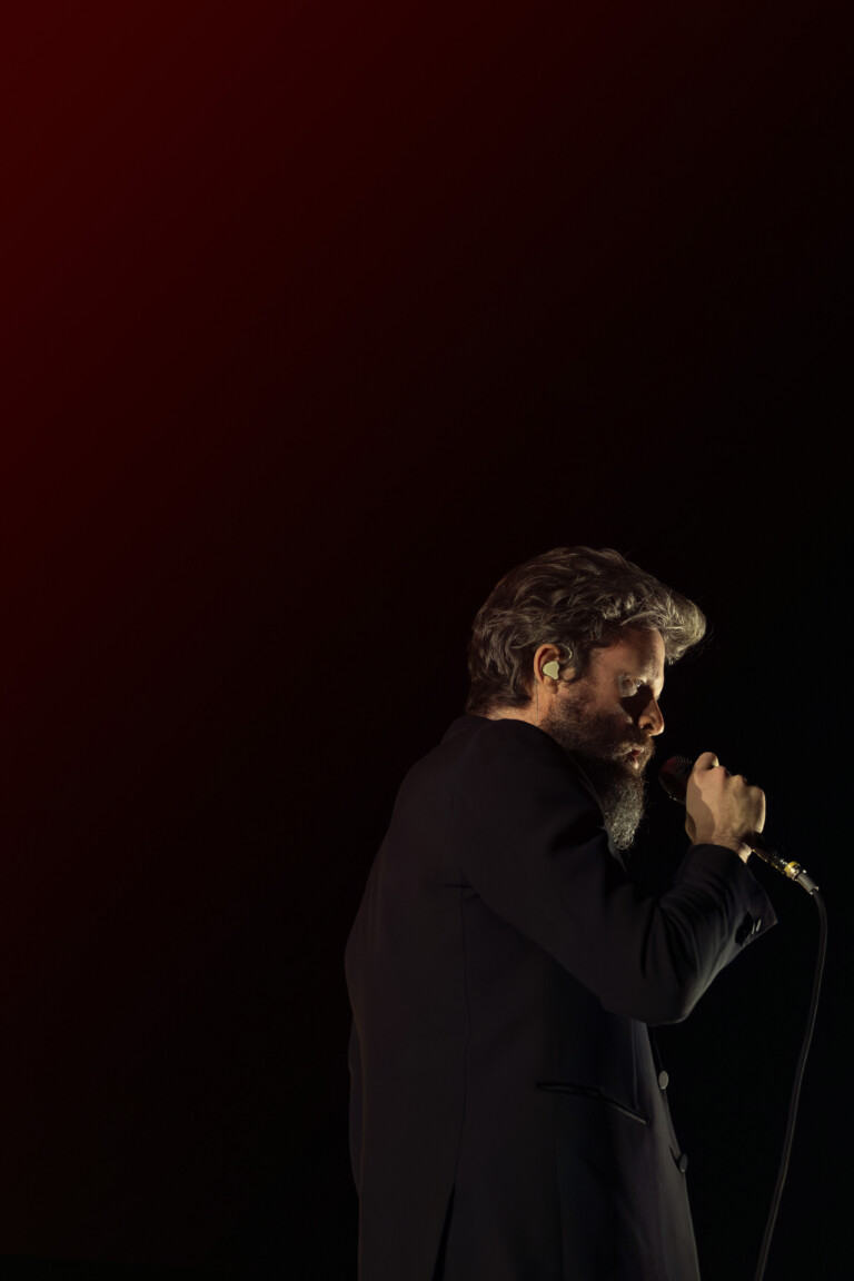 Father John Misty lowers his head in a medium shot while holding the microphone.