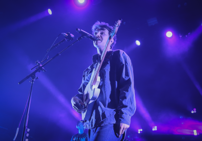 flipturn's lead singer, Dillon Basse, singing with his guitar onstage. Purple lighting overhead.