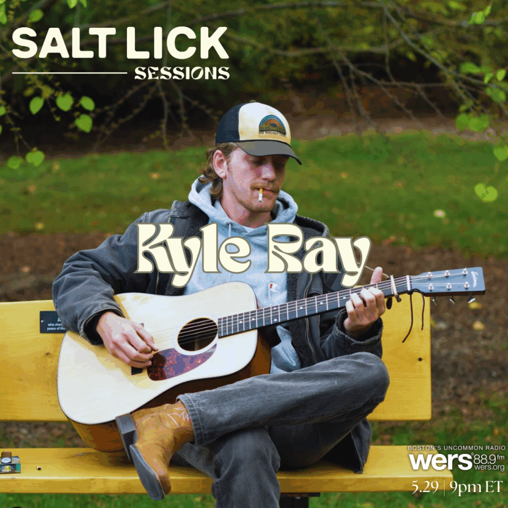 Kyle Ray sitting on a bench smoking a cigarette and playing a guitar. He's wearing jeans, boots, a hoodie with a work coat over it and a trucker style baseball hat.